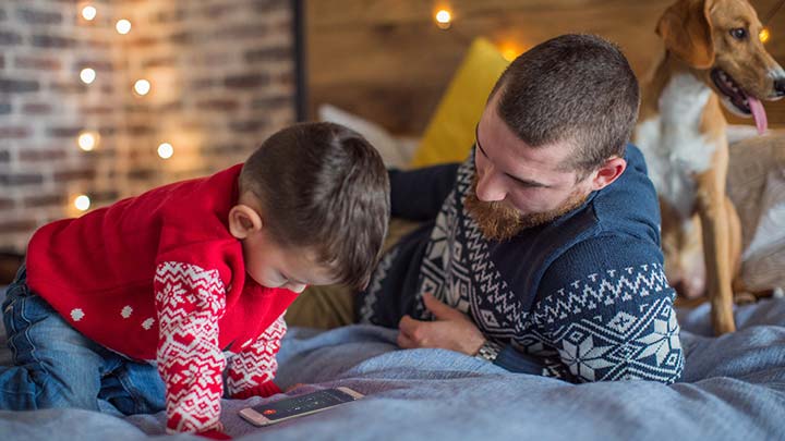 Father, son and pet dog sitting on bed with mobile phone to call Santa's Hotline with Christmas lights in background