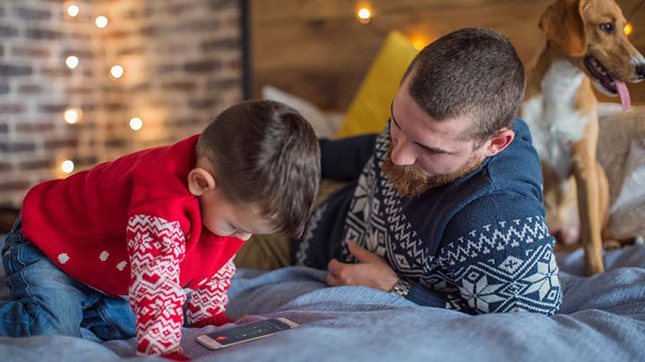 Far og sønn med hund som sitter på sengen med mobiltelefonen på vei til å ringe Julenissens Ønskelinje med julelys i bakgrunnen